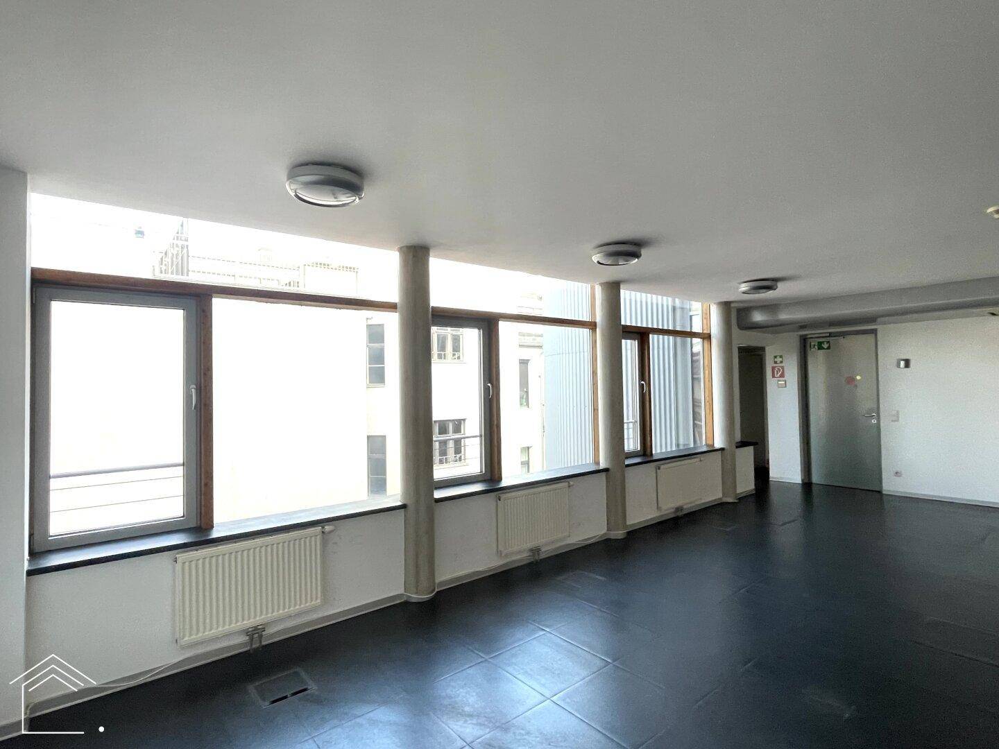 Langer Flur oder offener Bürobereich mit durchgehender Fensterfront und mehreren Betonsäulen.