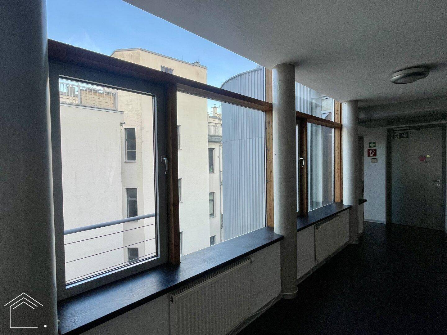 Heller Flur mit großen Fenstern, die einen Blick auf umliegende Gebäude bieten, und Betonsäulen.