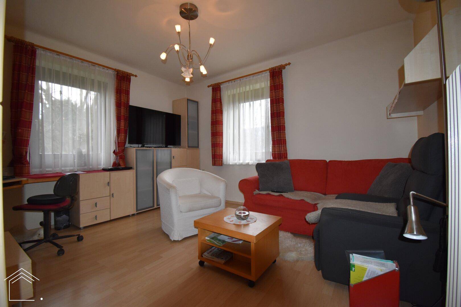 Helles Wohnzimmer mit rotem Sofa, Schreibtisch und Laminatboden, ideal für Entspannung und Arbeit.