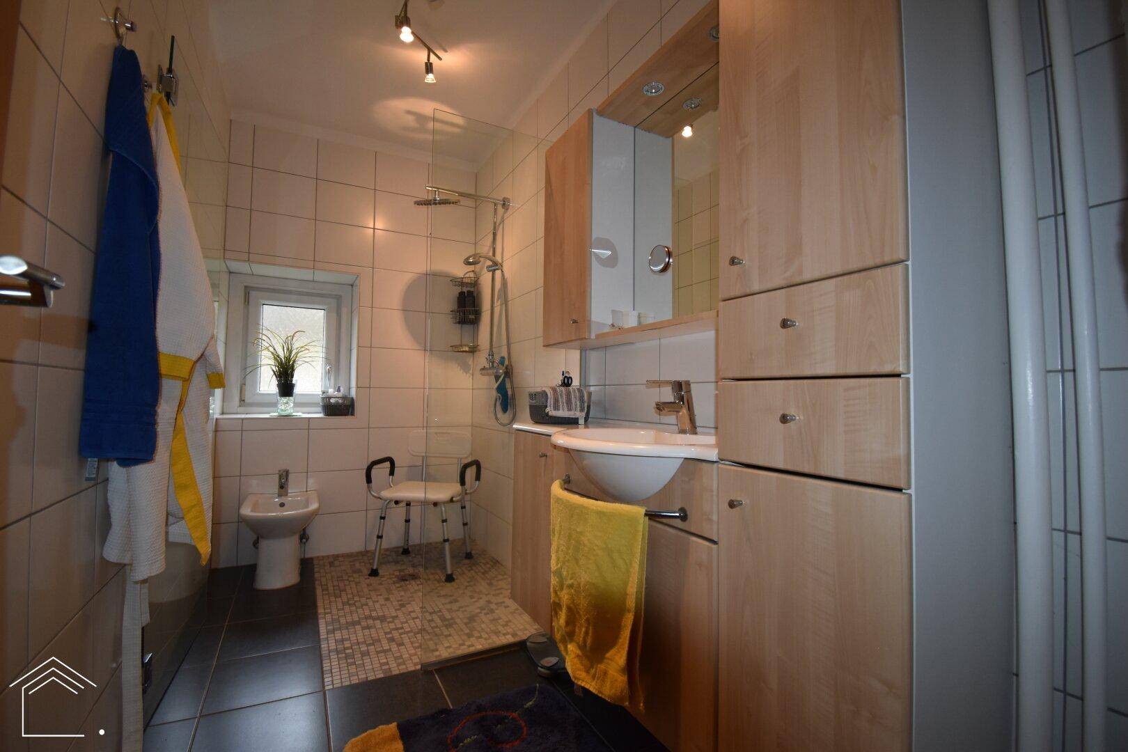 Helles Badezimmer mit Dusche, Bidet, Fenster und praktischen Holzschränken für Stauraum.