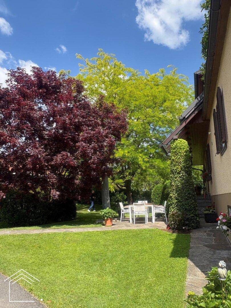 Sonniger Gartenbereich mit Rasen, bunten Bäumen und einer Sitzgruppe im Freien.