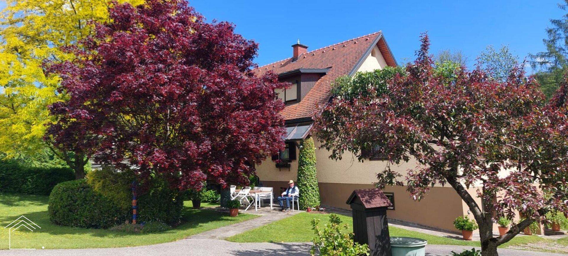 Charmantes Haus mit Ziegeldach, umgeben von einem grünen Garten und bunten Bäumen.