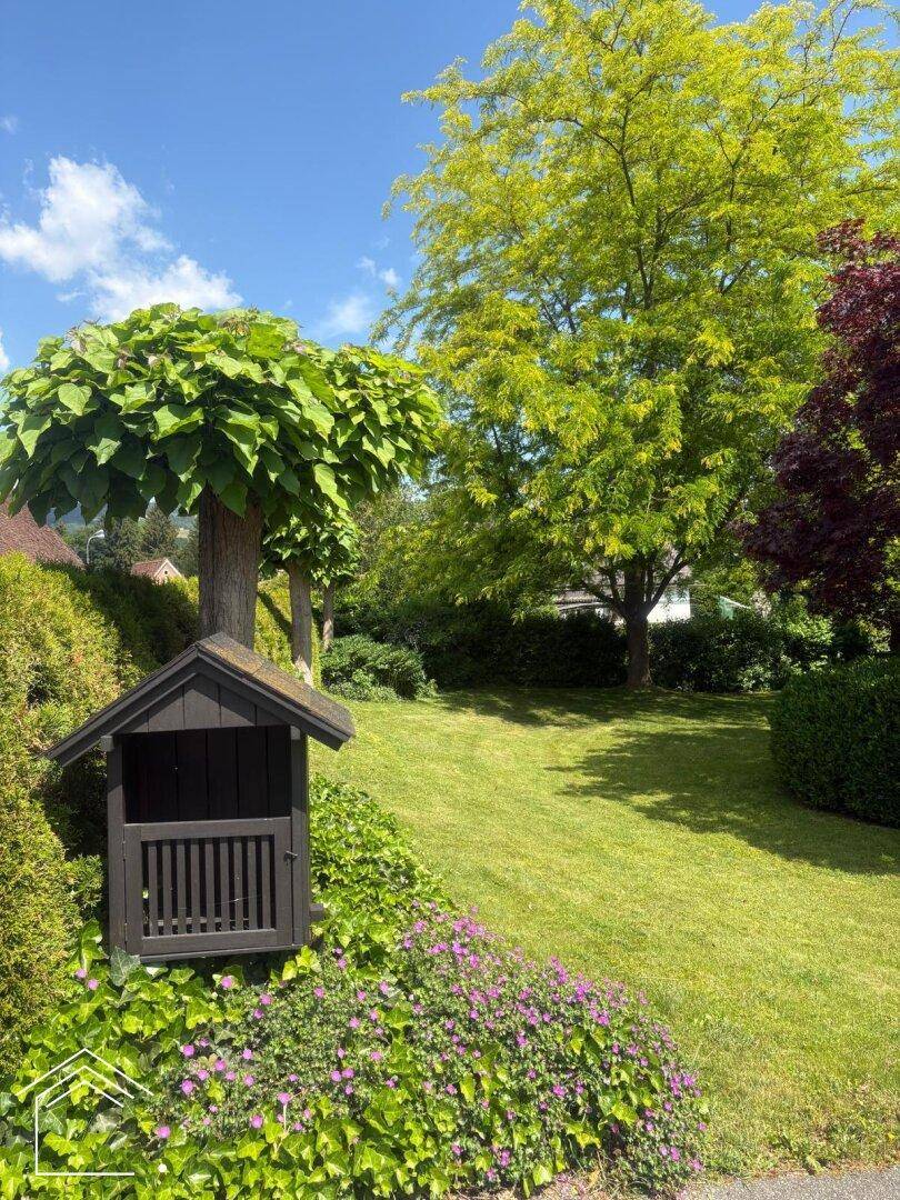 Gepflegter Garten mit einem kleinen Holzhaus, üppiger Bepflanzung und großen Bäumen.