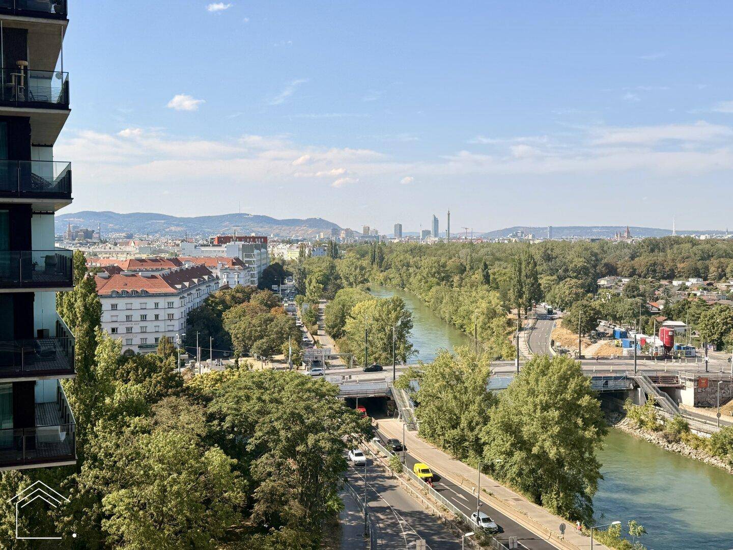 Panoramablick über Fluss, grüne Landschaften und die Stadt mit Bergen im Hintergrund.