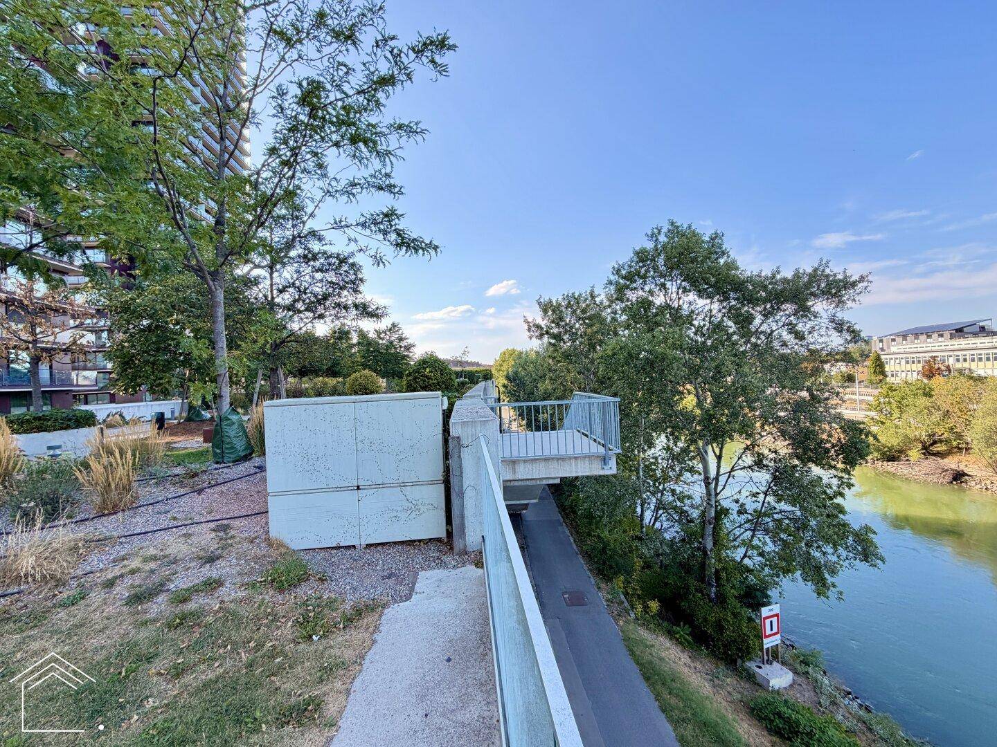 Uferpromenade mit Blick auf den Fluss und moderne Architektur im Hintergrund an einem sonnigen Tag.