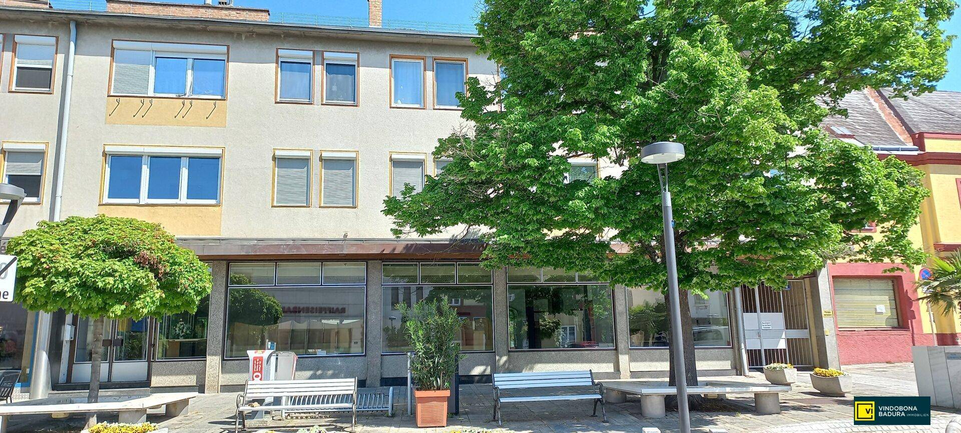 Mehrstöckiges Gebäude mit Geschäftsflächen im Erdgeschoss und Wohnungen darüber, umgeben von Bäumen und Bänken.