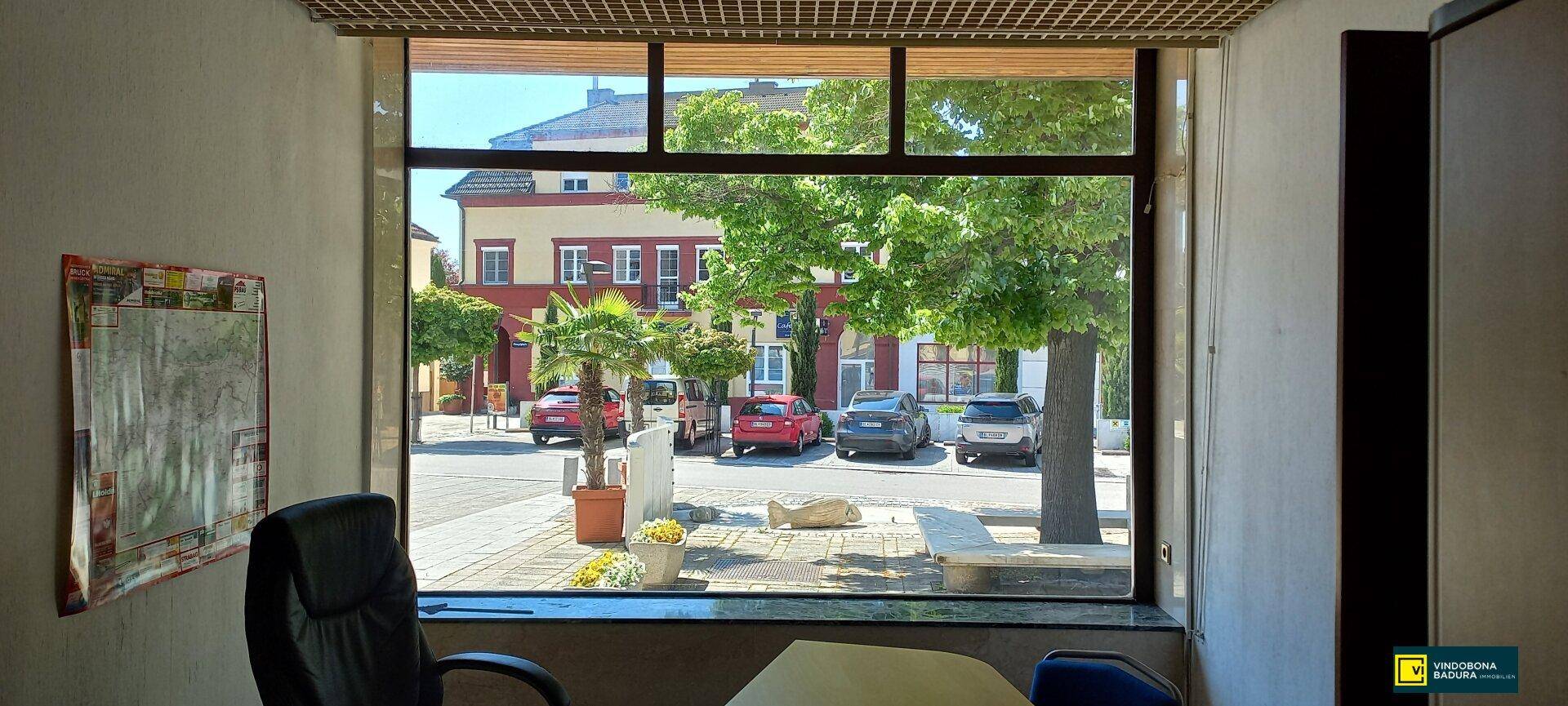 Blick aus einem Fenster auf eine belebte Straße mit parkenden Autos und gegenüberliegenden Gebäuden an einem sonnigen Tag.