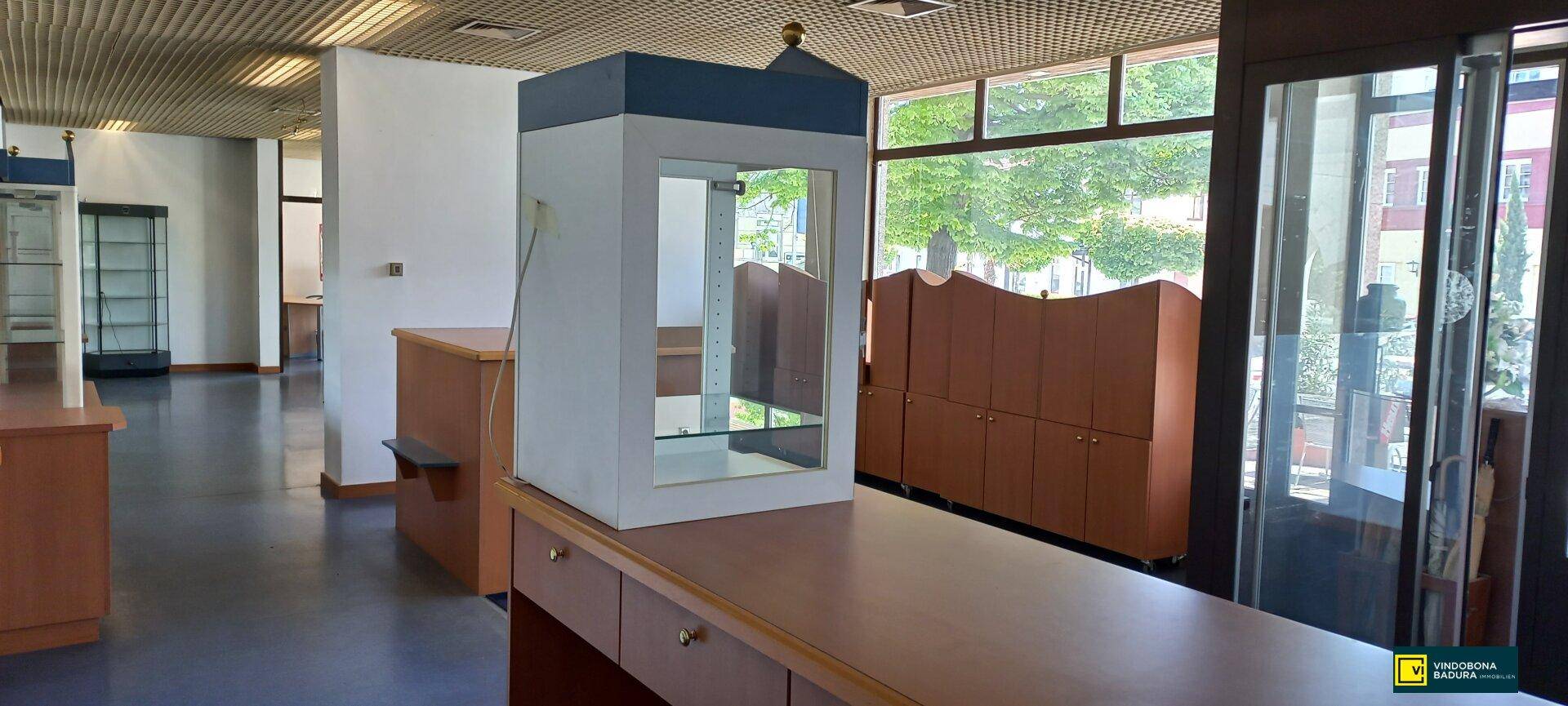 Verkaufsraum mit einer Theke, einer Vitrine und großen Fenstern, die viel Tageslicht hereinlassen und Holzmöbeln.