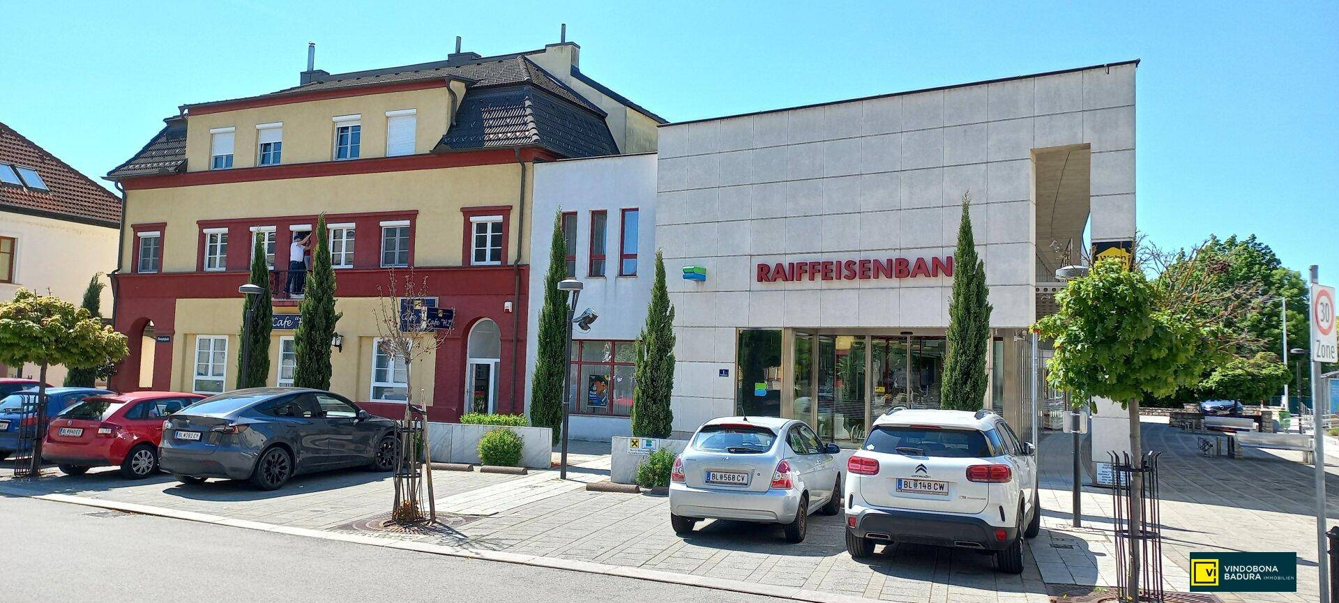 Straßenansicht mit geparkten Autos vor einer modernen Bank und einem angrenzenden Wohn- und Geschäftsgebäude.