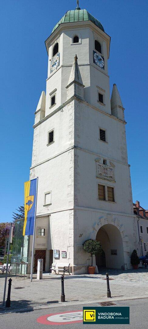 Hoher historischer Uhrturm mit grünem Kupferdach und einem Torbogen am Fuße des Turms unter blauem Himmel.