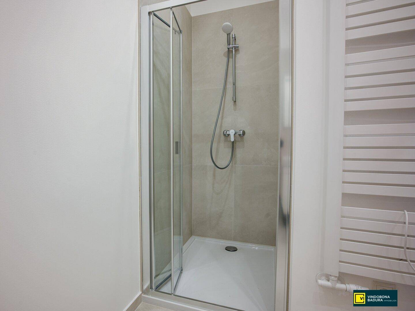Moderne Duschkabine mit Glasabtrennung und beigefarbenen Fliesen, ausgestattet mit einem Duschkopf.