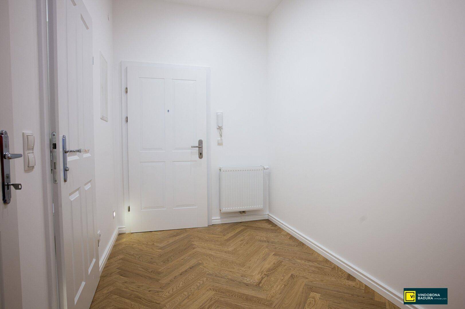 Heller Eingangsbereich mit Fischgrätparkettboden und weißer Eingangstür, die zu weiteren Räumen führt.
