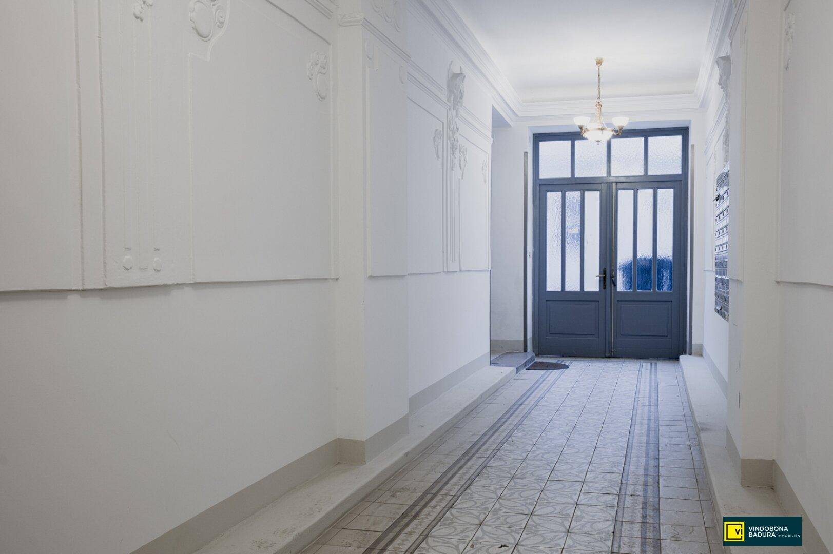 Geräumiger Eingangsbereich mit hohen Decken, Stuckelementen und einer großen Doppeltür zum Gebäudeausgang.