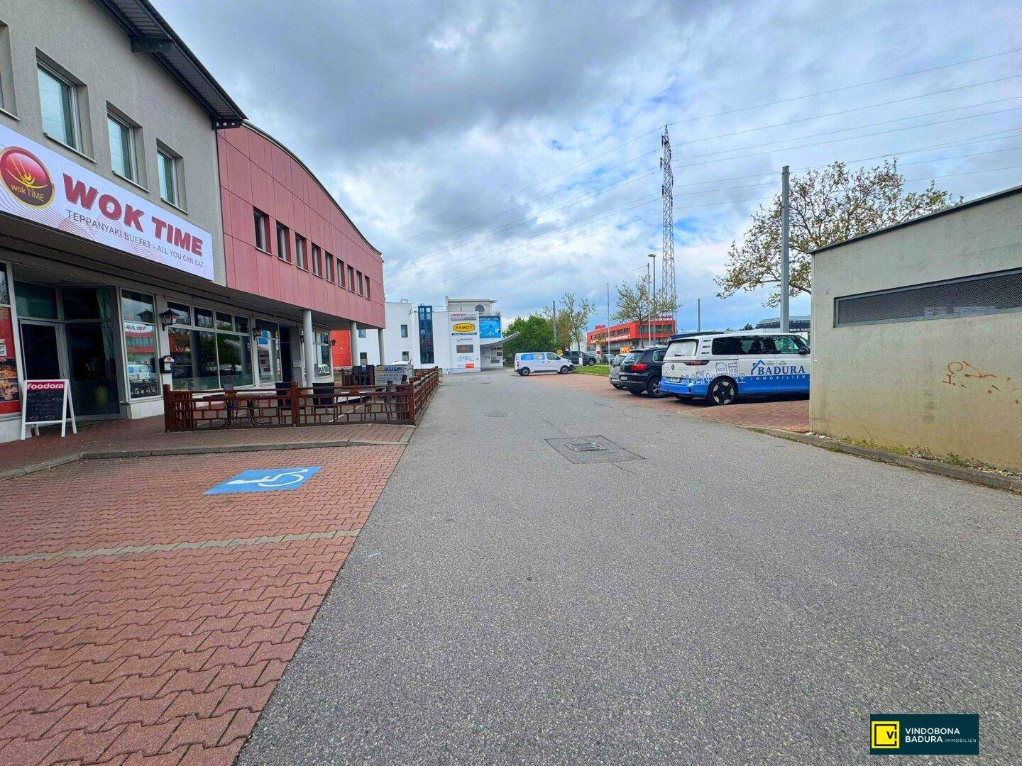 Seitenansicht eines Geschäftsgebäudes mit Außenbereich für Gastronomie und angrenzendem Parkplatz.