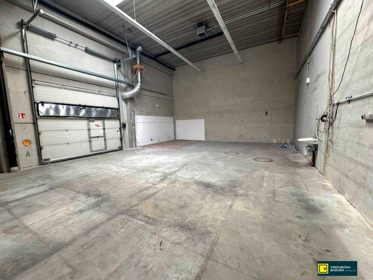 Leere Lagerhalle mit Betonboden, Rolltor und sichtbaren Versorgungsleitungen an den Wänden.