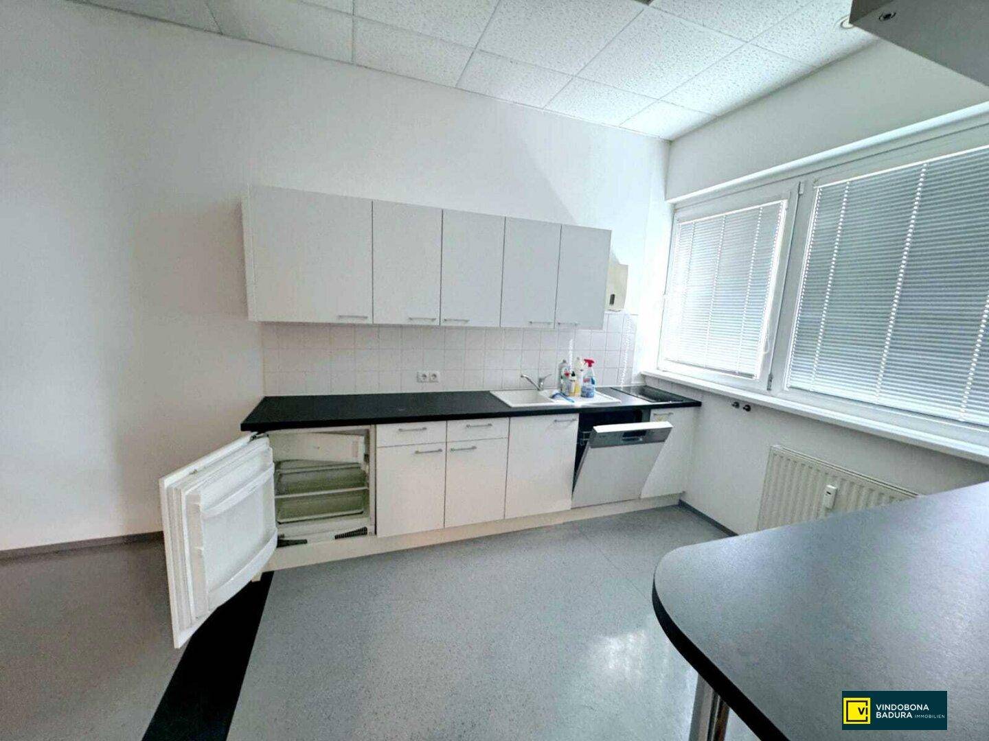 Funktionale Küchenzeile mit weißen Schränken, Spülmaschine, Kühlschrank und Fenster mit Jalousien.