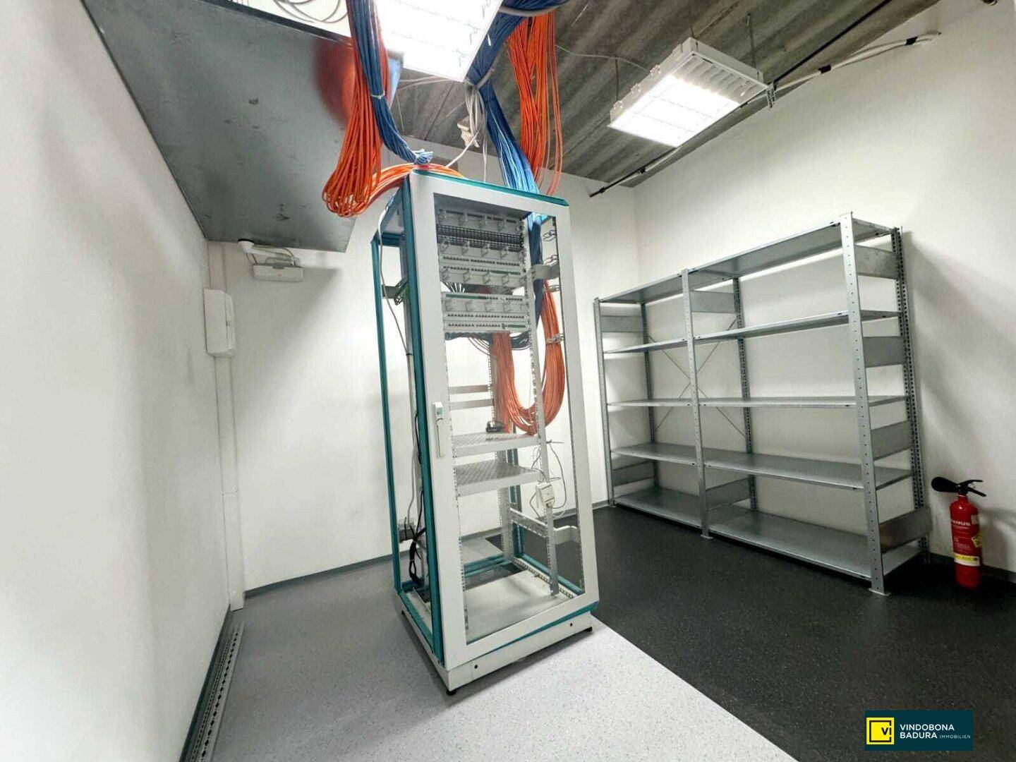 Technikraum mit Serverschrank, Kabelmanagement und Metallregalen für Lagerung oder Ausrüstung.