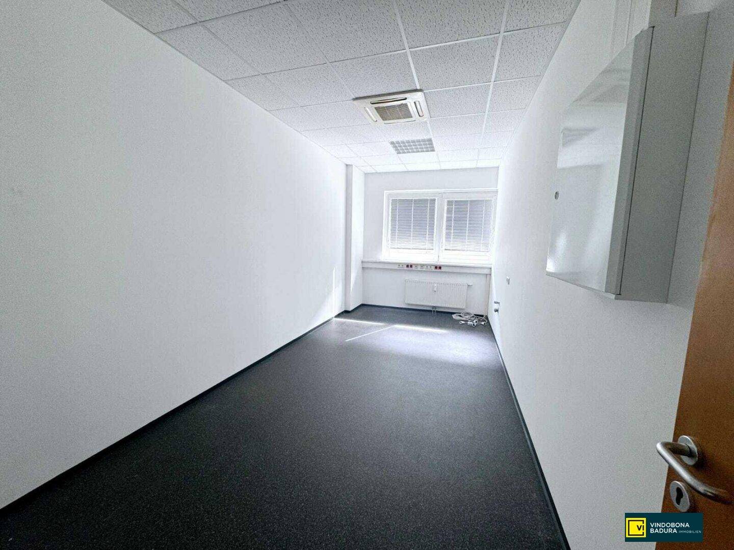 Kleineres Büro mit dunklem Bodenbelag, weißen Wänden und einem Fenster mit Jalousien am Ende des Raumes.