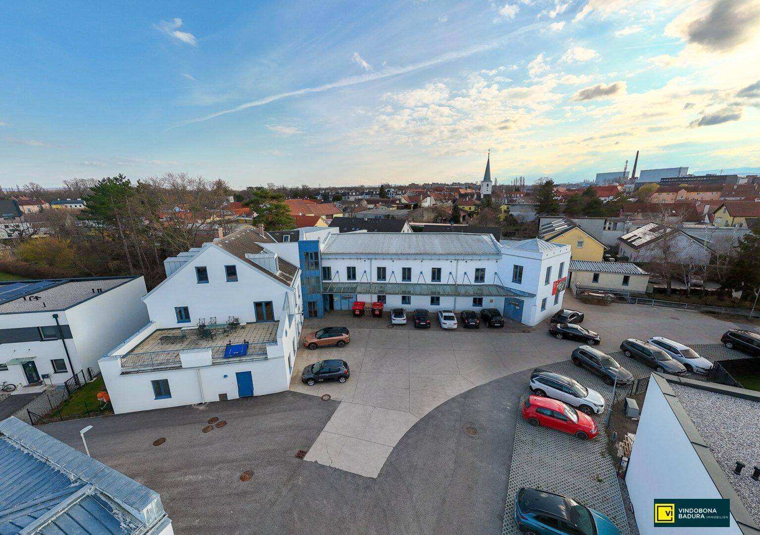 Panoramablick auf den Gebäudekomplex mit Parkplatz und der umliegenden Stadtlandschaft im Hintergrund.