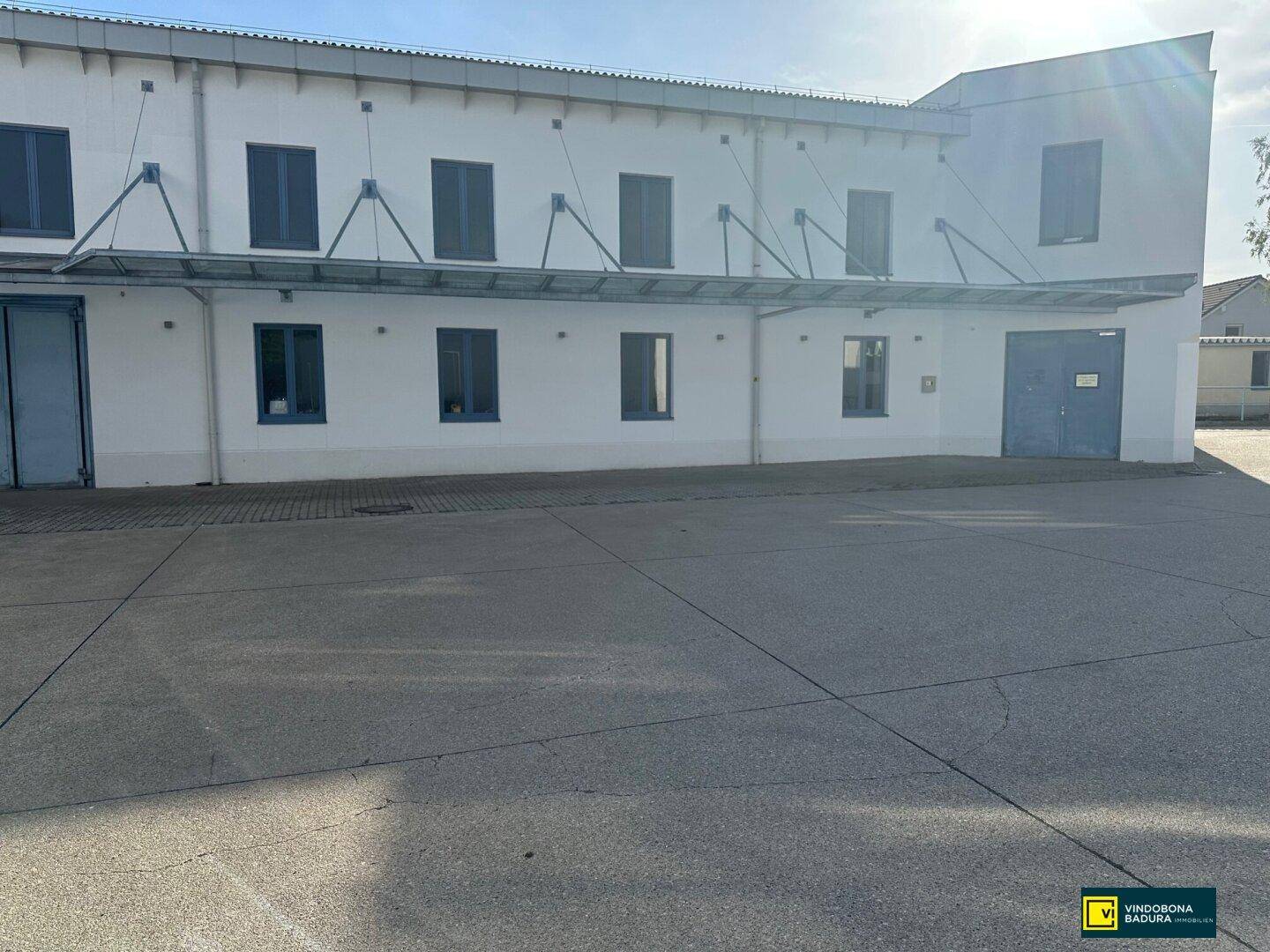Ansicht der Gebäudefassade mit großen Fenstern und einem weitläufigen gepflasterten Vorplatz.