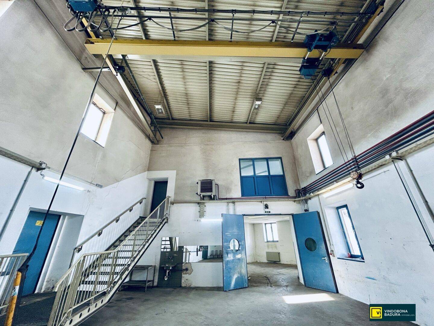 Großer Industrie- oder Lagerraum mit Krananlage, Treppe und blauen Türen.