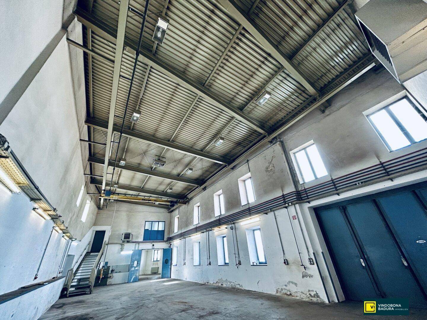 Weitläufiger Industrie- oder Lagerraum mit hohen Decken, Krananlage und vielen Fenstern.