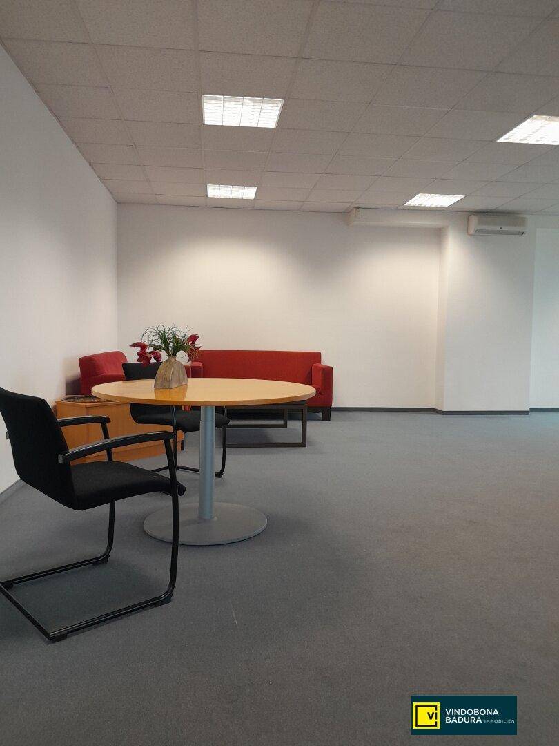 Gemütlicher Loungebereich im Büro mit rotem Sofa, rundem Tisch und Stühlen auf grauem Teppichboden.