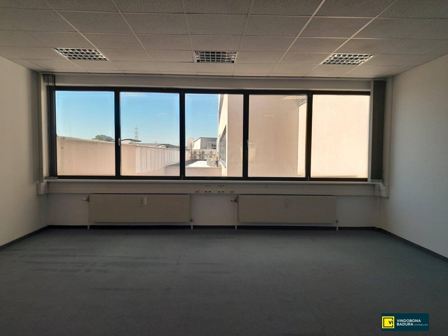 Leeres Büro mit breitem Fenster, das einen Blick auf umliegende Gebäude und den Himmel freigibt.