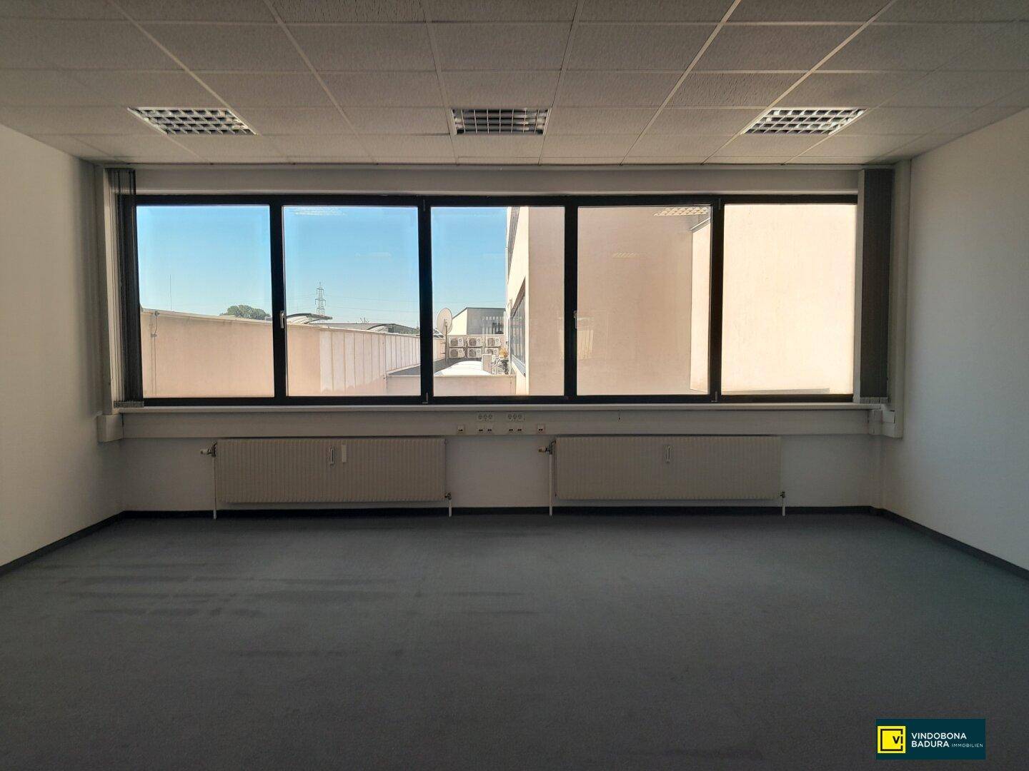 Leeres Büro mit breitem Fenster, das einen Blick auf umliegende Gebäude und den Himmel freigibt.