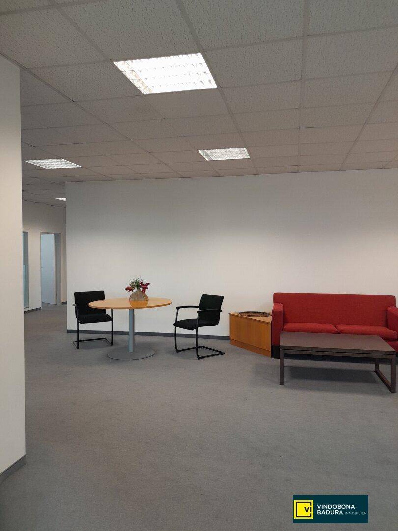 Gemütlicher Loungebereich im Büro mit rotem Sofa, rundem Tisch und Stühlen auf grauem Teppichboden.