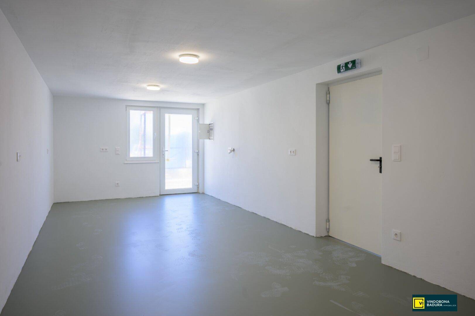 Leerer Abstellraum mit grauem Boden, weißen Wänden und einer Tür nach draußen.