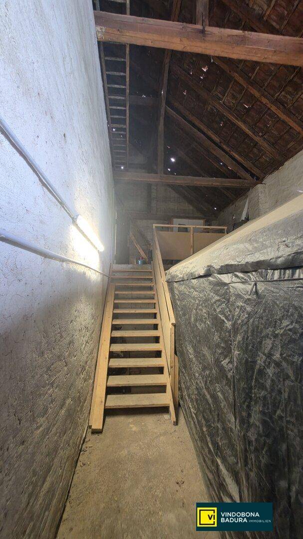 Schmaler Innenraum mit Holztreppe, die zu einem Dachboden mit sichtbaren Holzbalken führt.