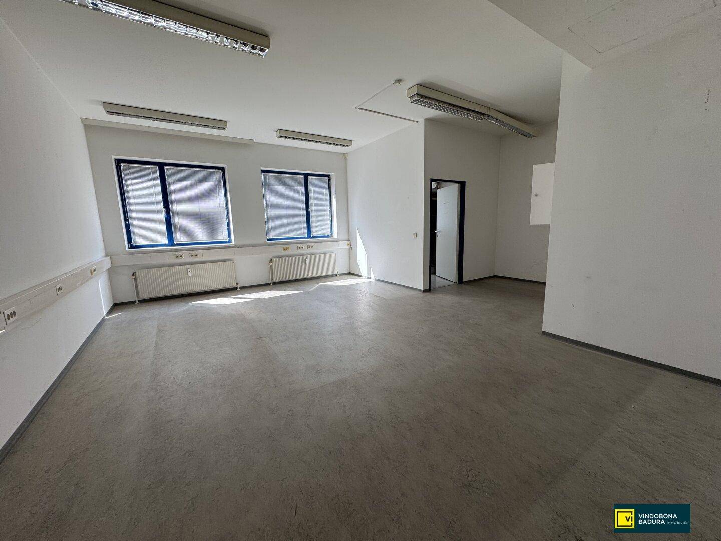 Leerer Büroraum mit Zugangstür, Fenstern und funktionalem Bodenbelag, bereit zur Einrichtung.