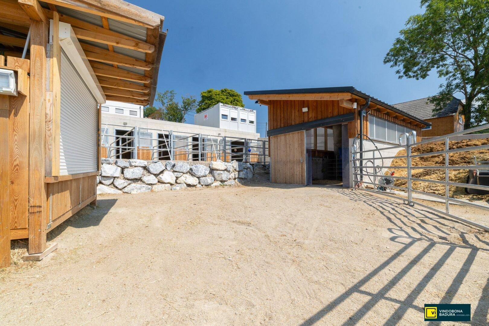 Ansicht des Pferdestalls mit Holzelementen, Steinfundament und weitläufigem Außenbereich bei Sonnenschein.