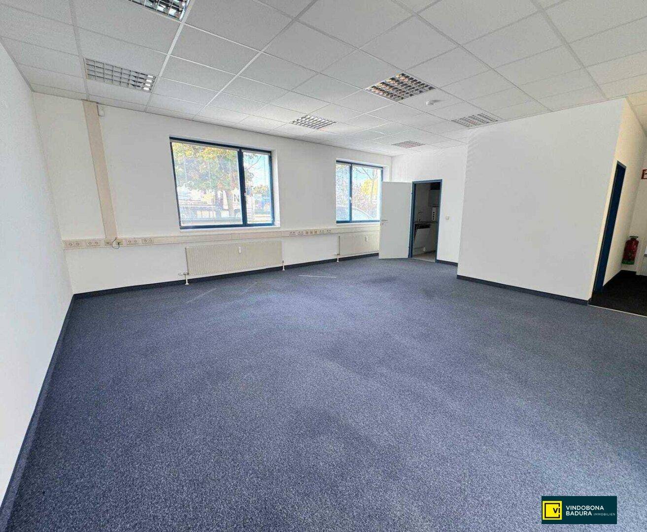 Leeres Büro mit Teppichboden, weißen Wänden und Zugang zu einem weiteren Raum.