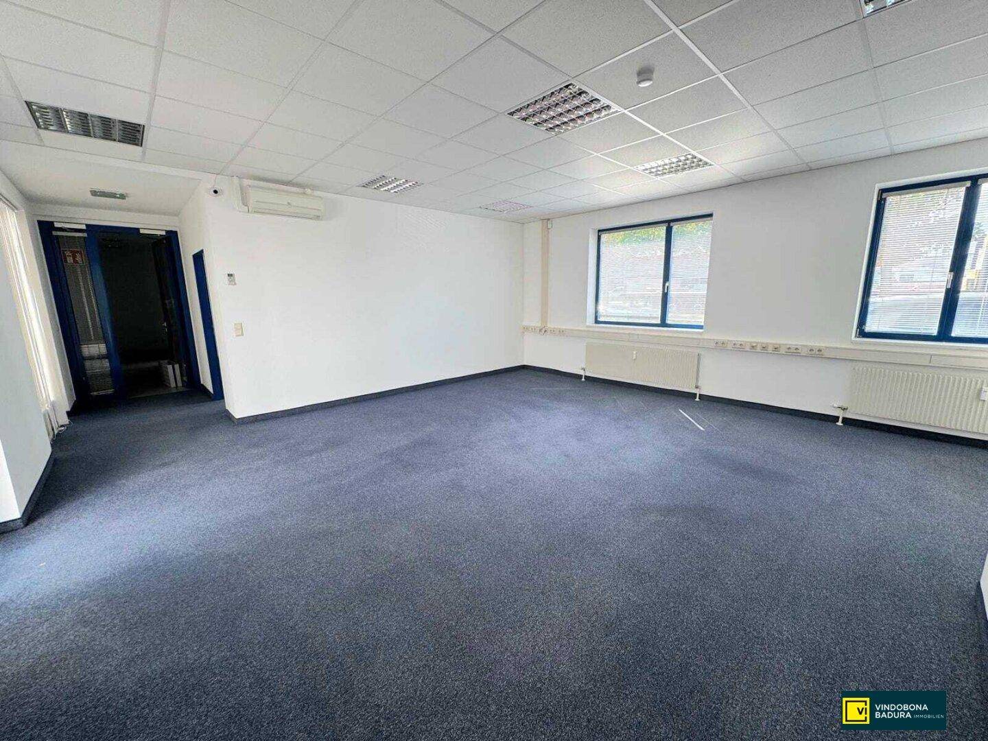 Helles Büro mit Teppichboden, Klimaanlage und Fenstern mit Blick nach draußen.