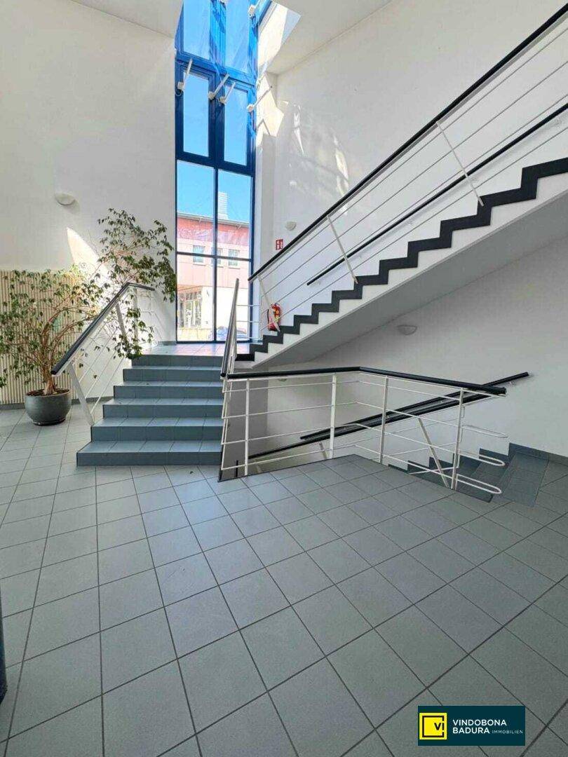 Heller Treppenaufgang mit großen Fenstern, Fliesenboden und Metallgeländer.