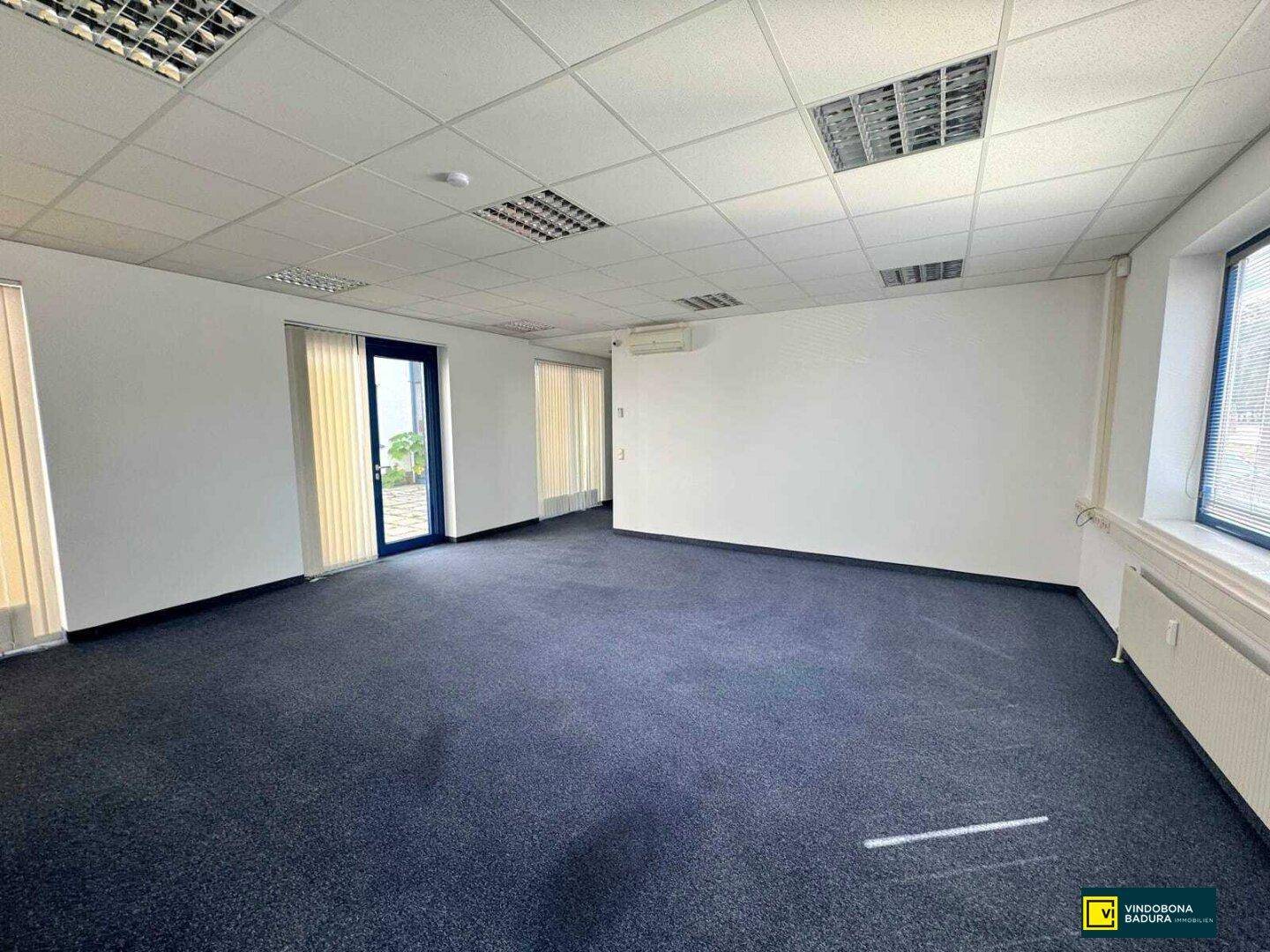 Geräumiges Büro mit Teppichboden, weißen Wänden und Zugang zu einem Balkon oder einer Terrasse.