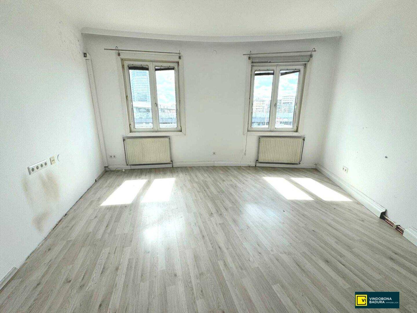 Geräumiges Zimmer mit hellem Laminatboden und zwei Fenstern, die viel Tageslicht hereinlassen.