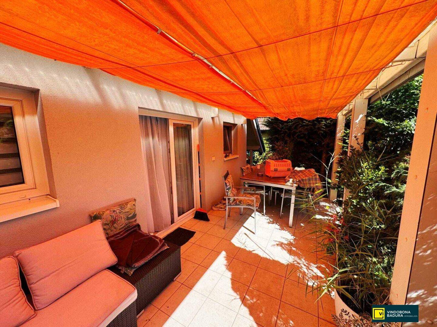 Überdachte Terrasse mit Fliesenboden, Gartenmöbeln und einem orangefarbenen Sonnenschutz.