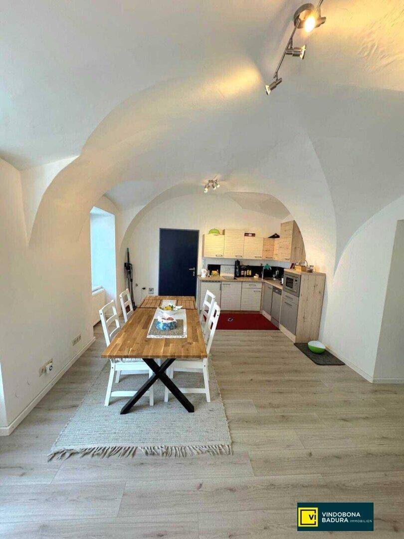 Gemütlicher Essbereich mit Holztisch, Stühlen und einer kleinen Küche unter Gewölbedecken.