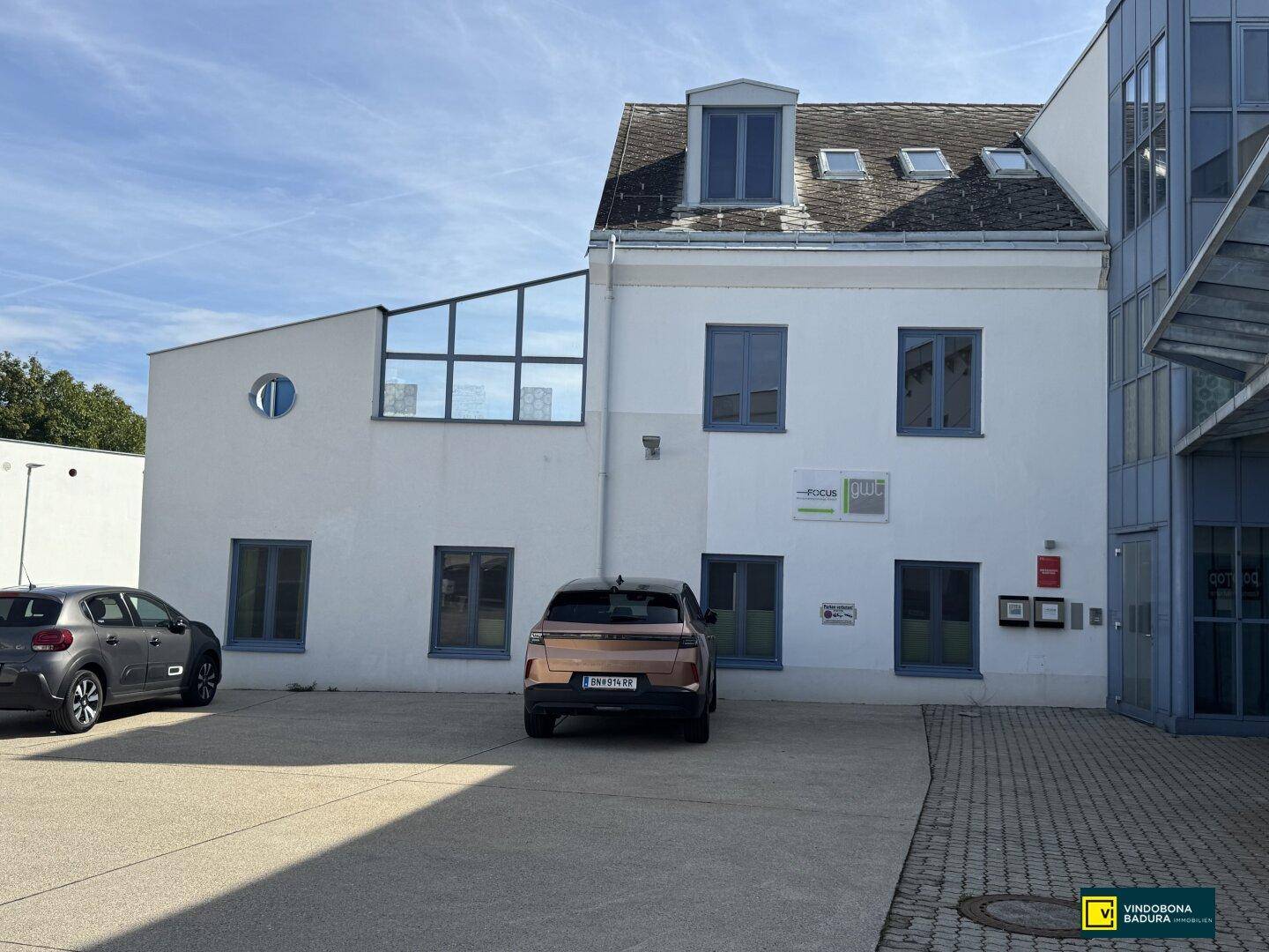 Fassade eines modernen Gebäudes mit Parkplätzen und großen Fenstern unter blauem Himmel.