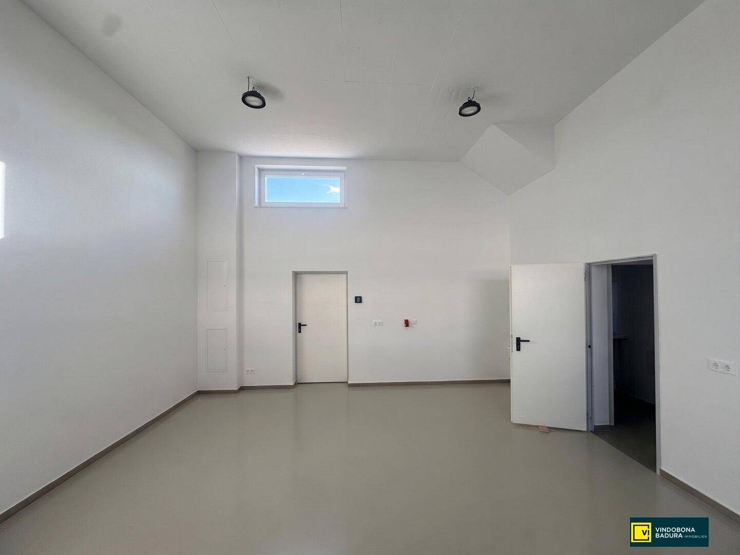 Heller, leerer Gewerberaum mit hohen Decken, weißen Wänden und einem kleinen Fenster oben.
