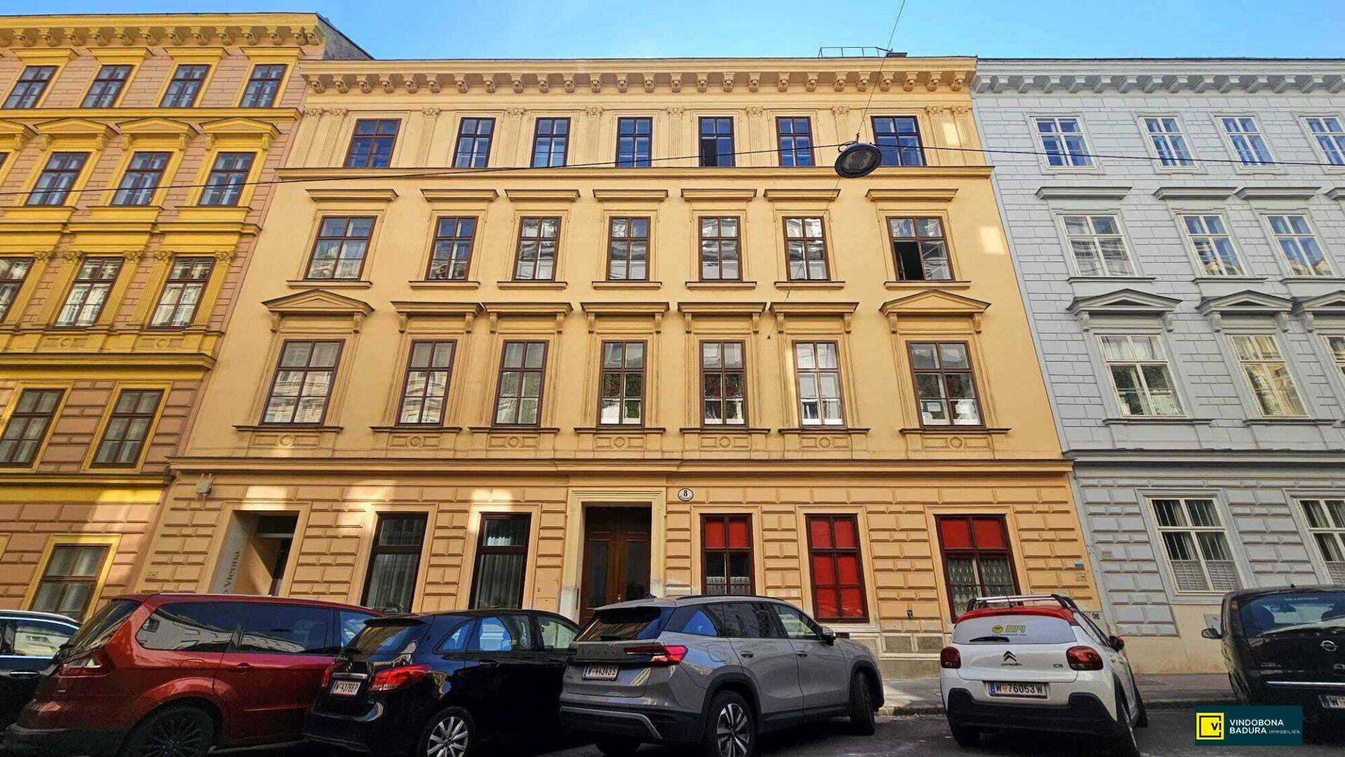 Historische Fassade eines Wohngebäudes mit vielen Fenstern und parkenden Autos davor.