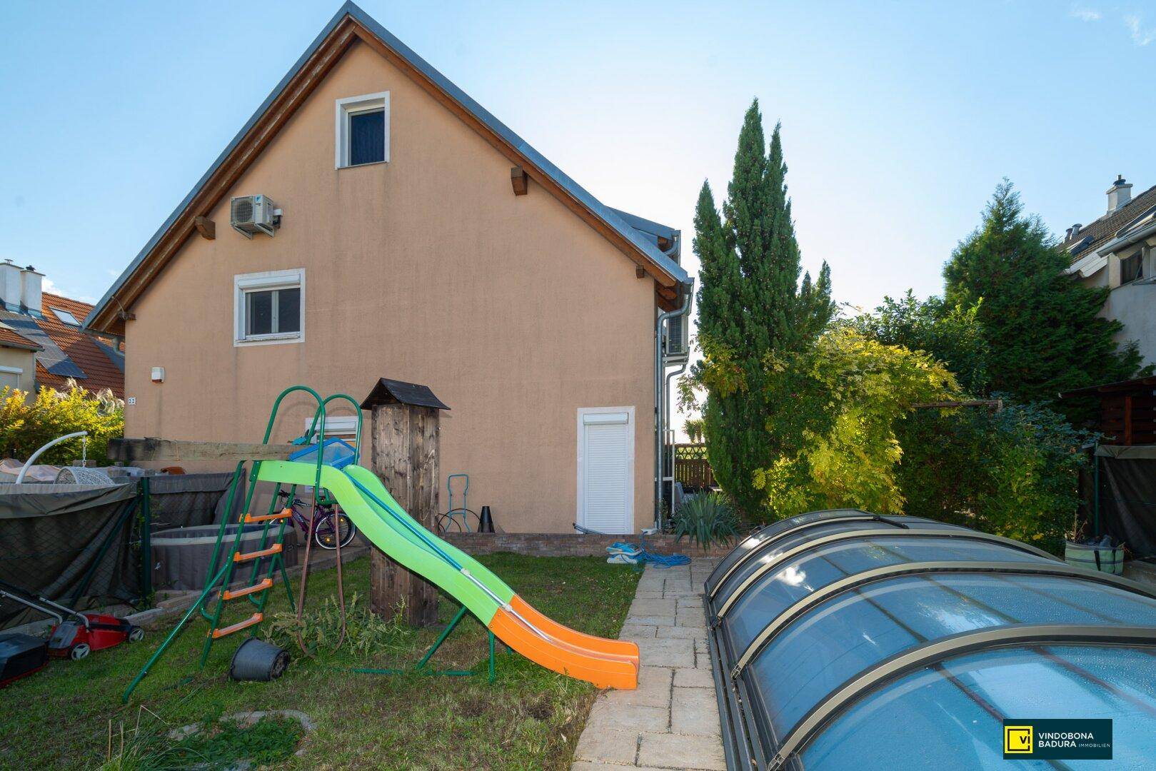 Gartenansicht des Hauses mit Poolüberdachung, Kinderrutsche und Rasenfläche an einem sonnigen Tag.
