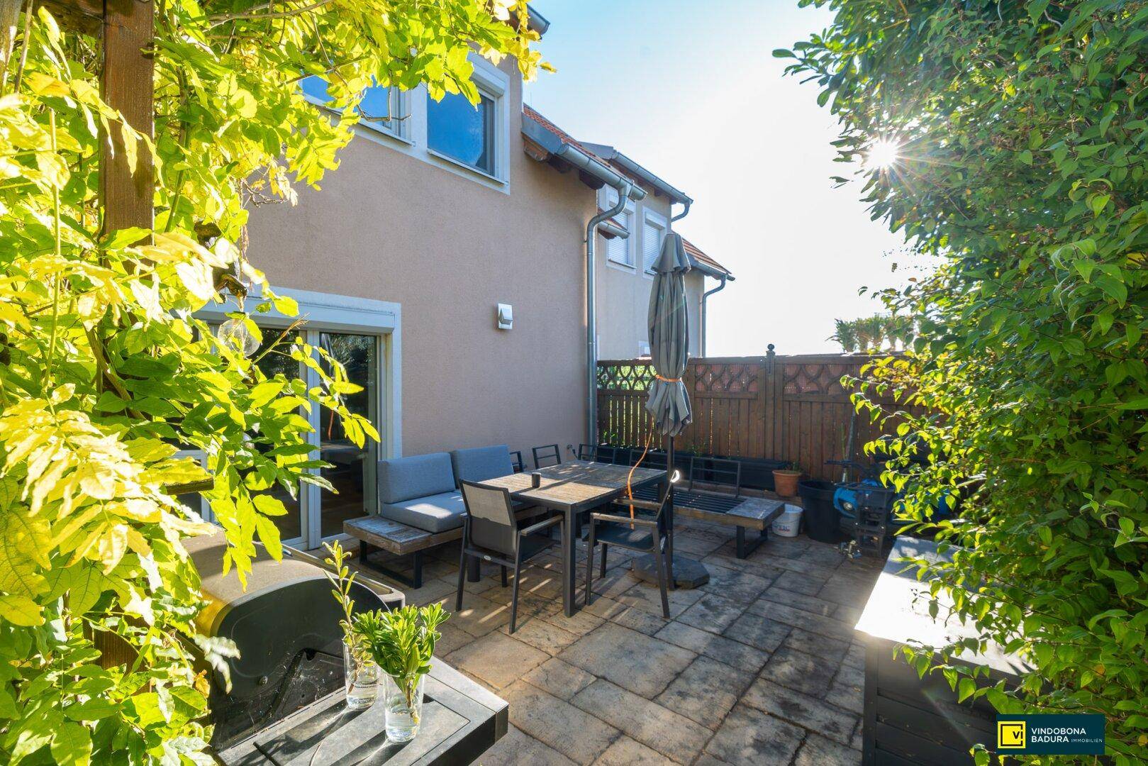 Gemütliche Terrasse mit Esstisch, Sitzgelegenheiten und Grillbereich, umgeben von üppigem Grün.
