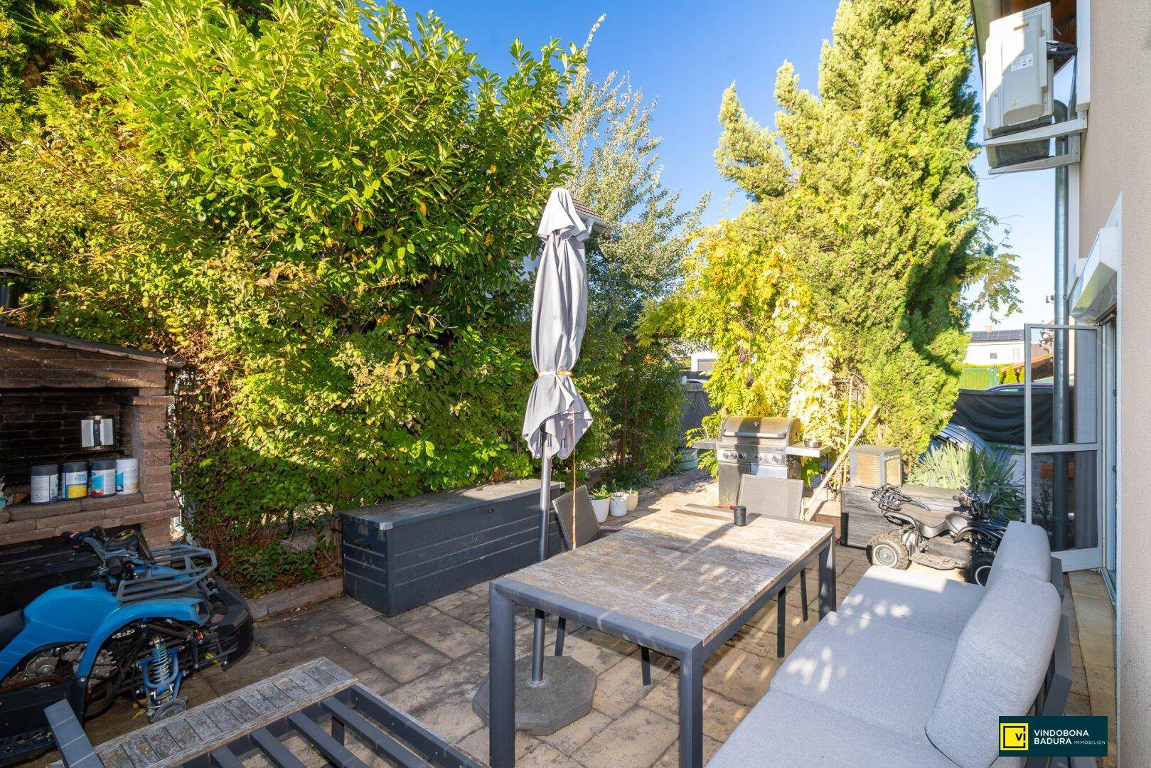Geräumige Terrasse mit Sitzmöbeln, Sonnenschirm und Grill, umgeben von hohen Hecken für Privatsphäre.
