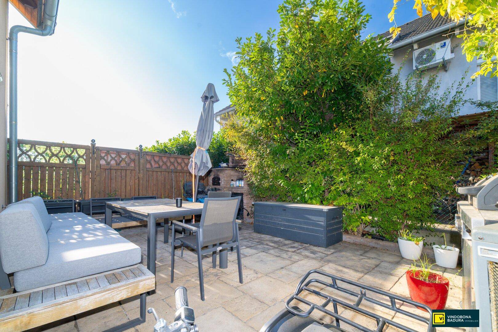 Sonniger Terrassenbereich mit Sitzmöbeln, Grill und Außenofen, umgeben von üppigem Grün.