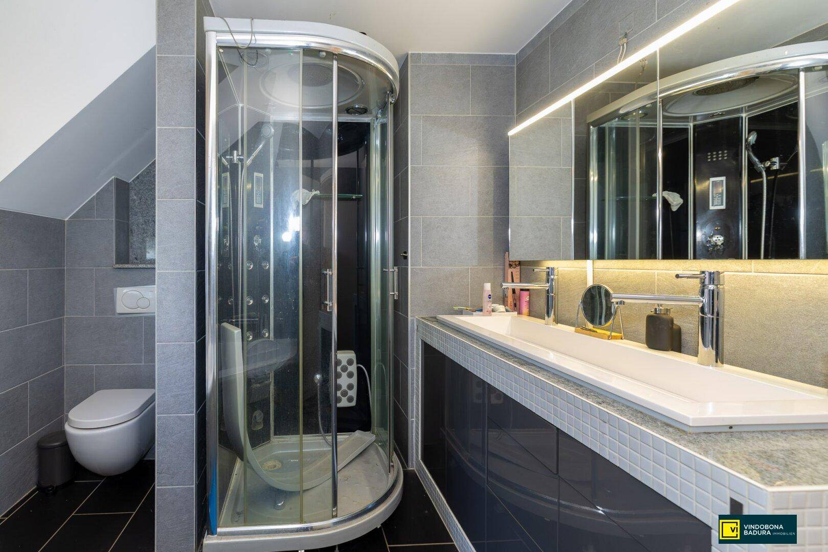 Elegantes Badezimmer mit Doppelwaschbecken, moderner Duschkabine und grauen Fliesen.