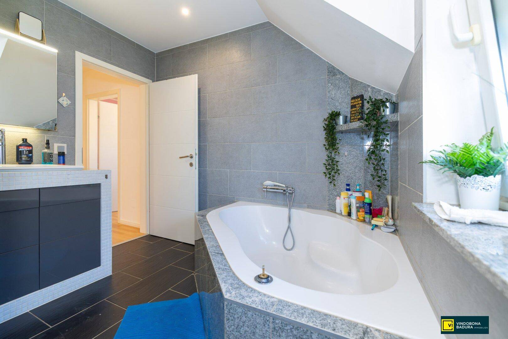 Geräumiges Badezimmer mit Eckbadewanne, Fenster und modernen grauen Fliesen, hell und einladend.